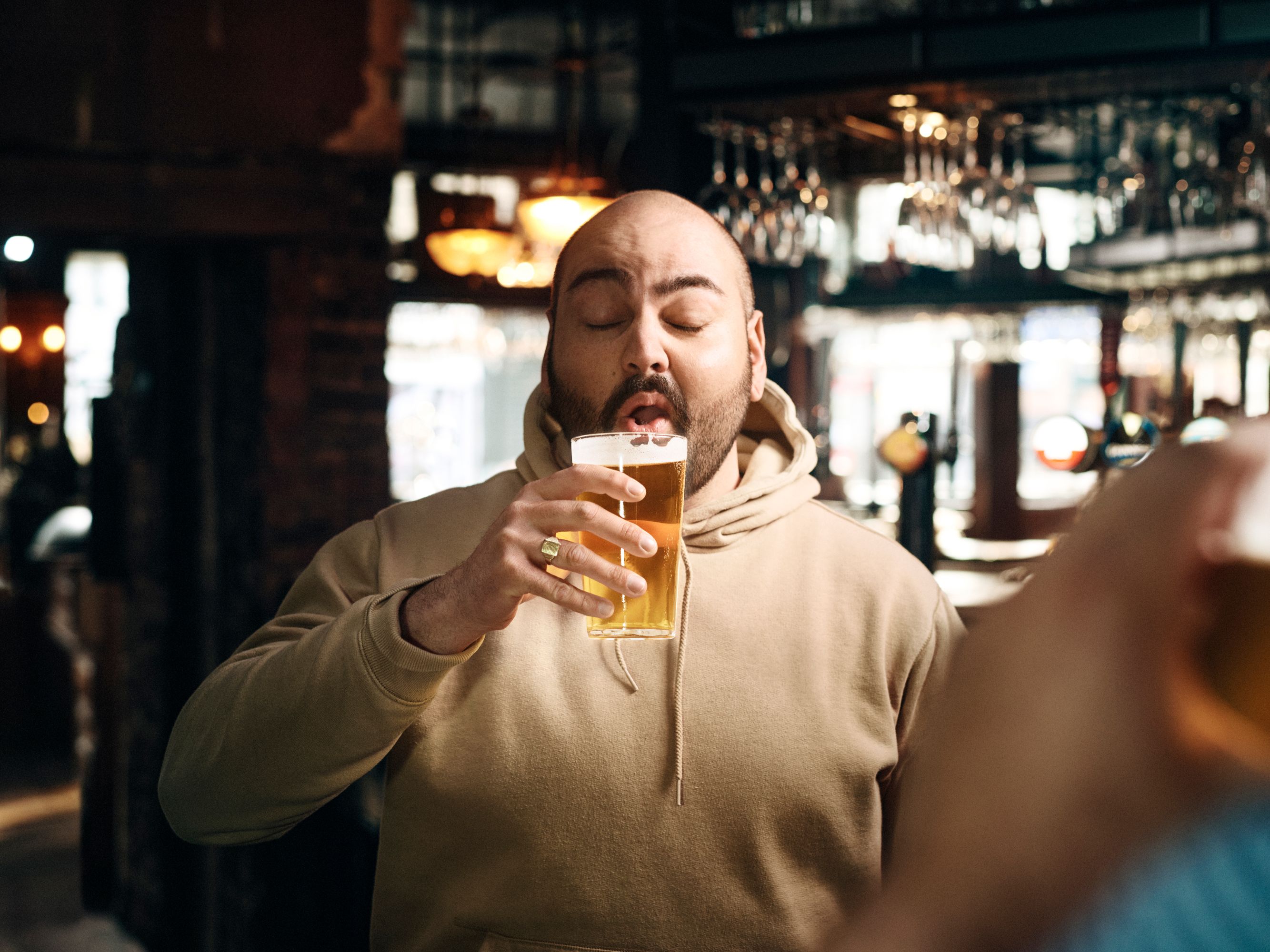 A man in a beige hoodie is drinking a beer in a dimly lit bar, with a blurred background of people and drinks.