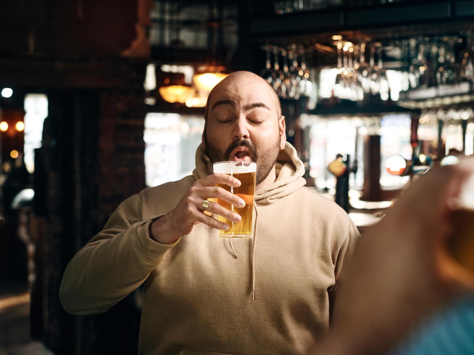 A man in a beige hoodie is drinking a beer in a dimly lit bar, with a blurred background of people and drinks.