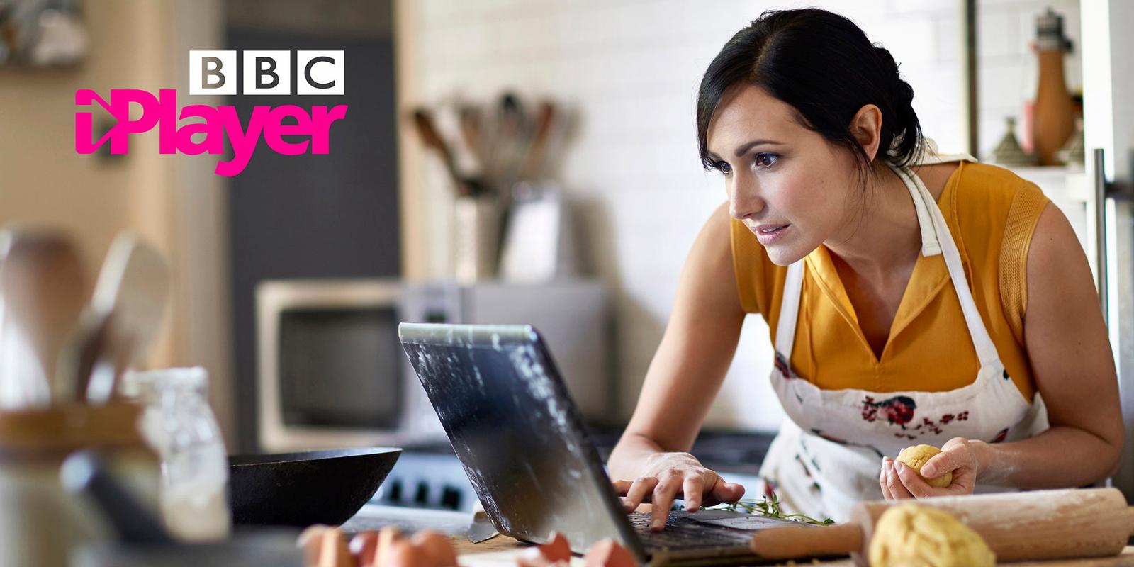 A woman in a yellow top and white apron is using a laptop in a kitchen, with a frying pan and other kitchen tools in the background.