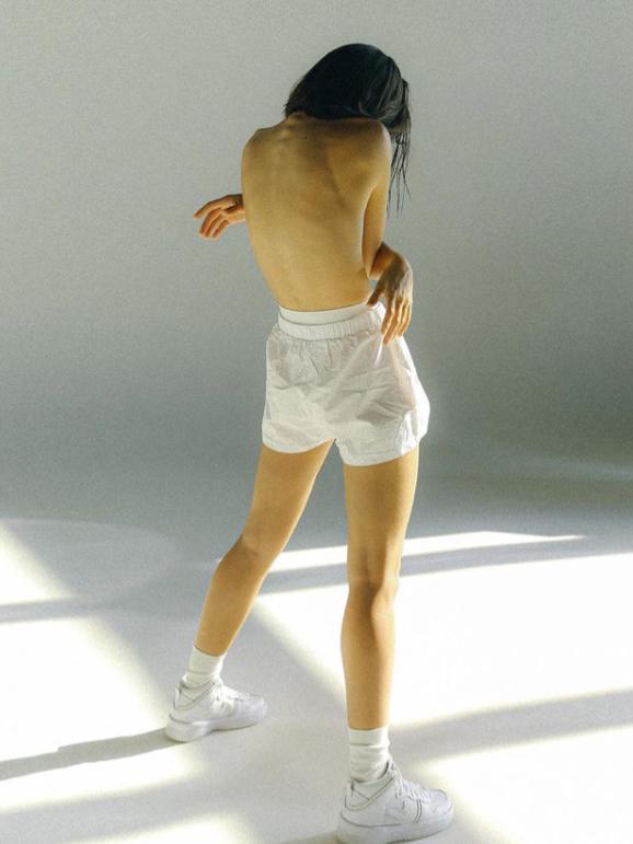 A woman with long, dark hair and pale skin, wearing only white socks and standing in a dimly lit room with her back to the camera, exposing her bare torso.