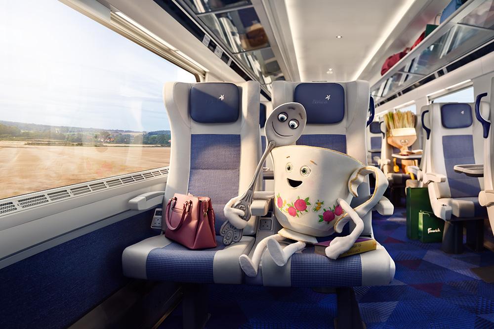 A train car interior with a large window showing a rural landscape, with a white and blue seat in the foreground and a red and white seat in the background.