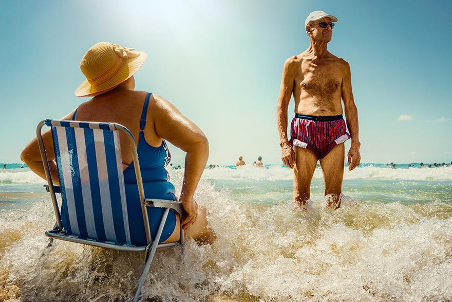 A woman in a blue swimsuit and a man in red and white striped swim trunks are standing in the ocean, with the woman sitting in a beach chair and the man standing in the water.