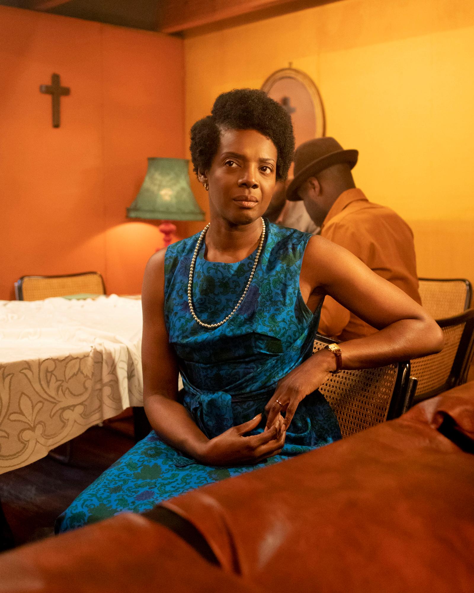 A woman in a blue dress with a floral pattern sits in a dimly lit room with a table and chairs, a cross on the wall behind her.