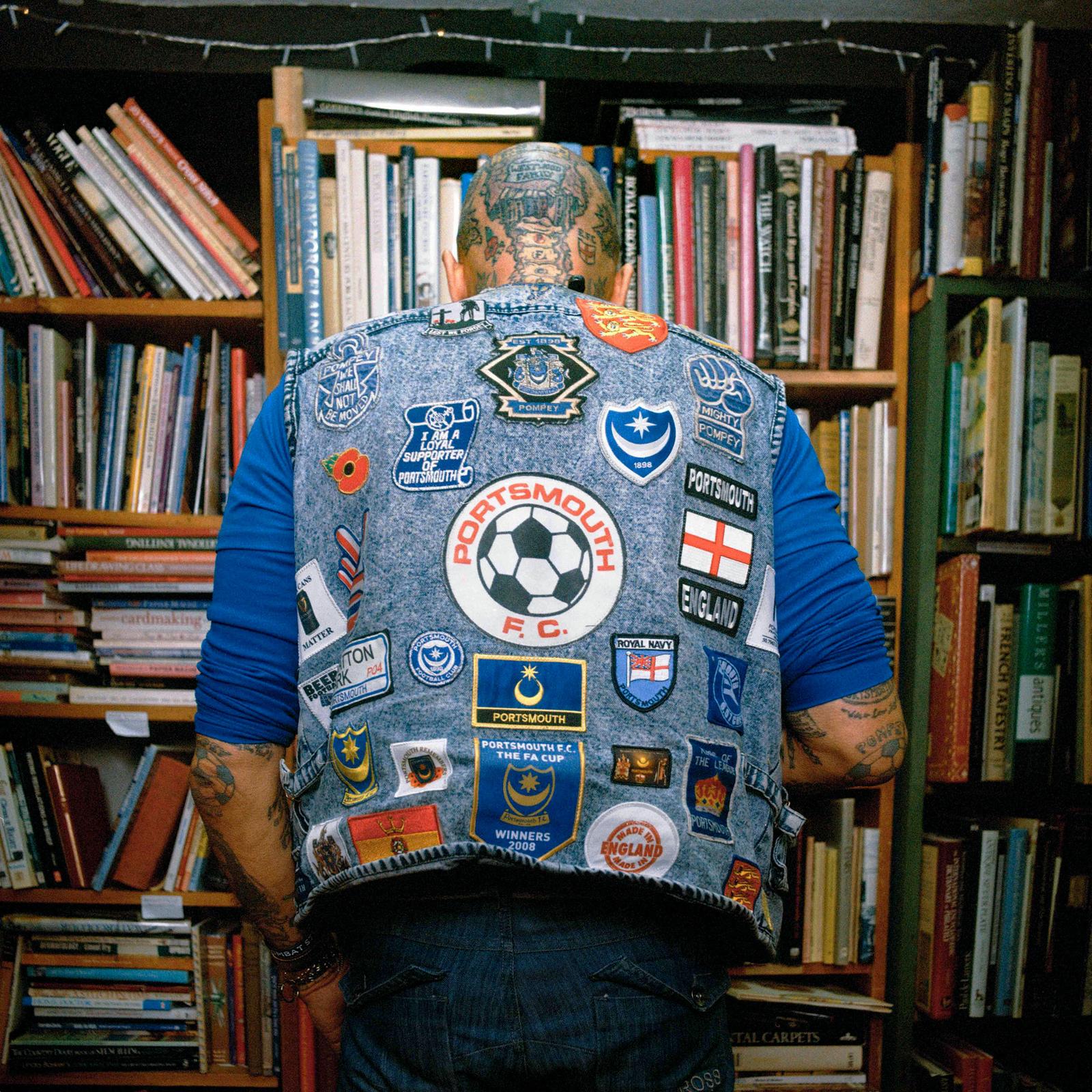 A man stands in front of a bookshelf wearing a denim jacket with patches and pins.