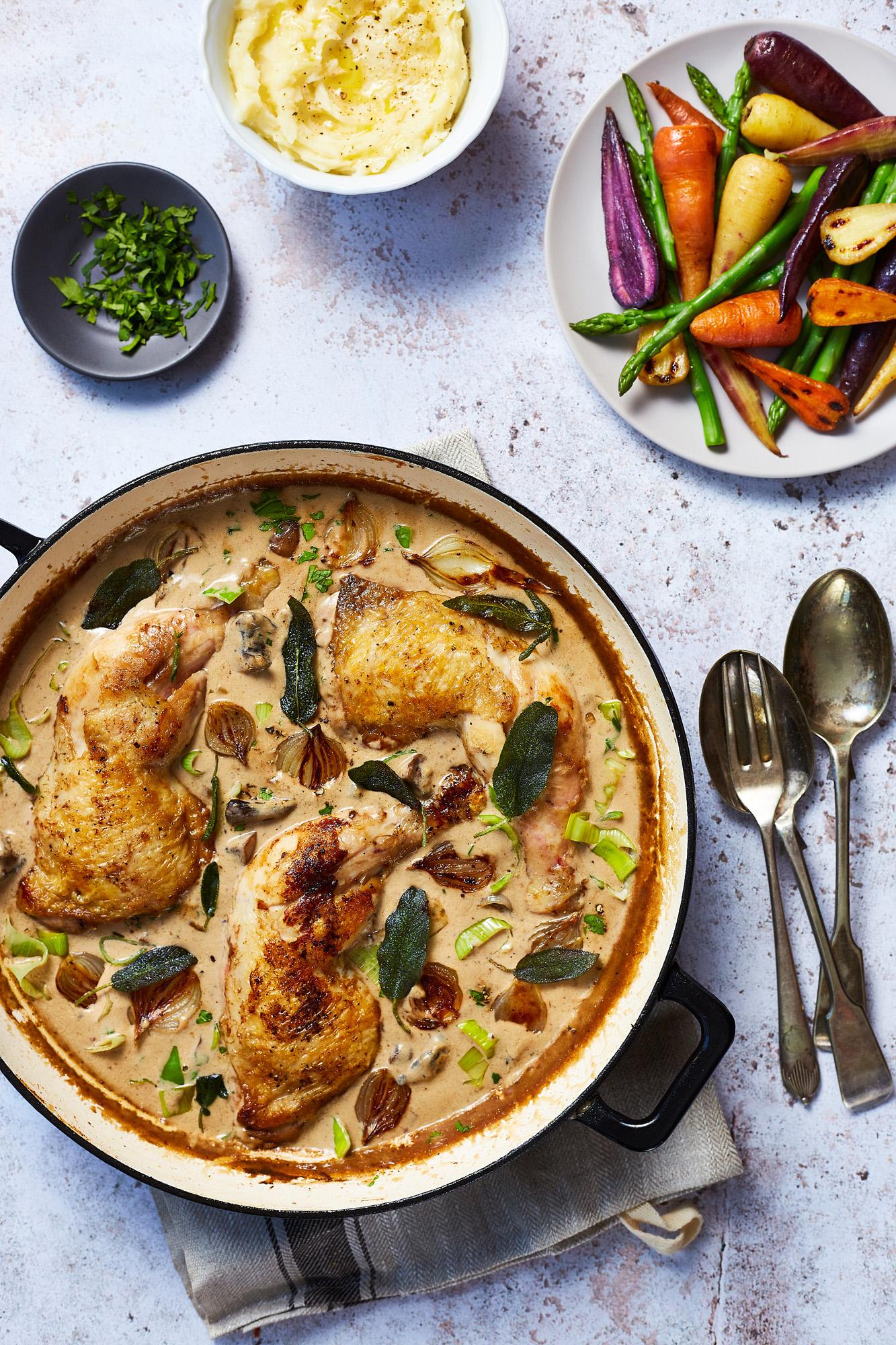 A large, golden-brown chicken dish in a creamy sauce, garnished with fresh herbs, served with a side of roasted vegetables and a bowl of mashed potatoes.