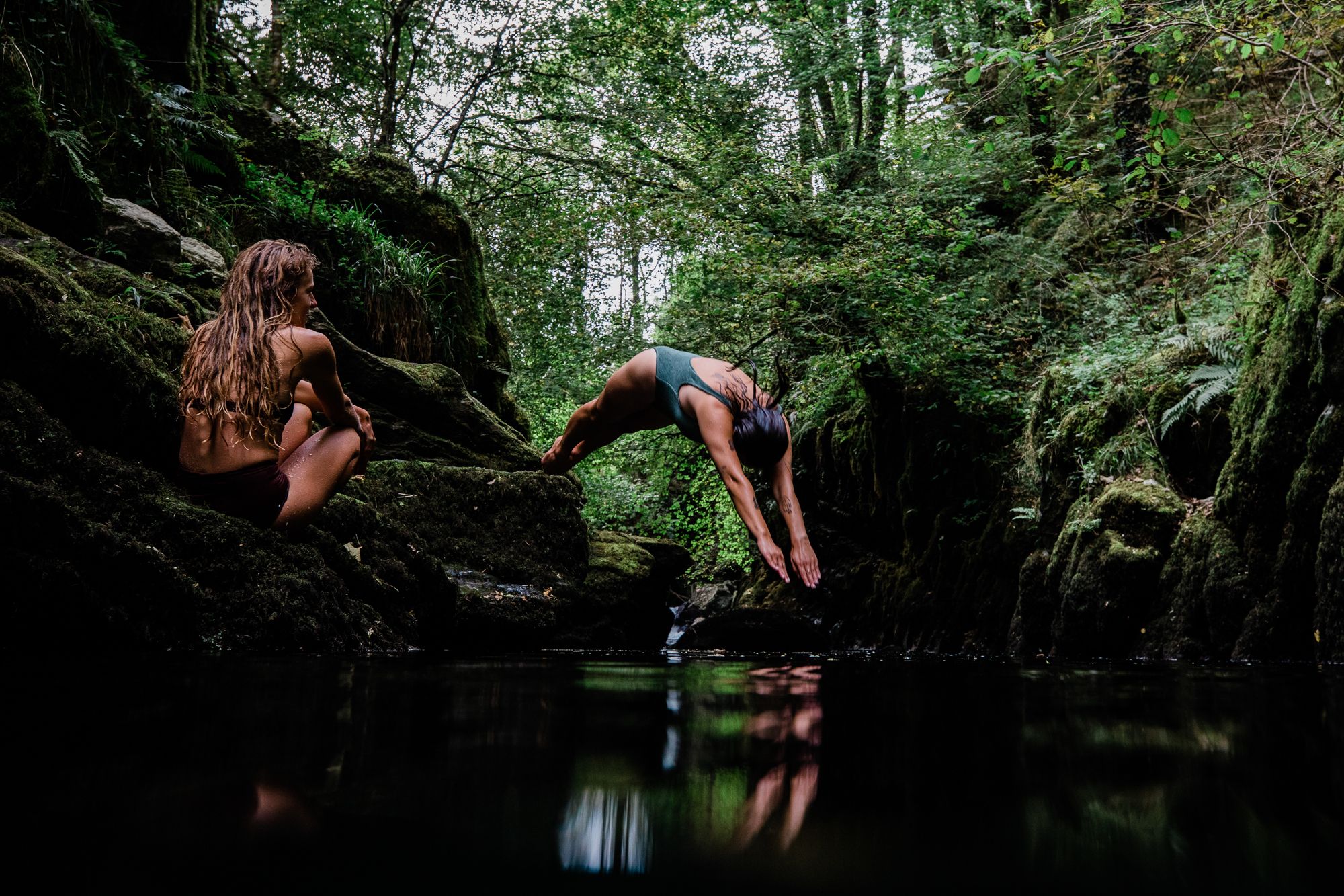 Two women in a forested area, one in a green swimsuit and one in a black swimsuit, both in a yoga pose near a body of water.