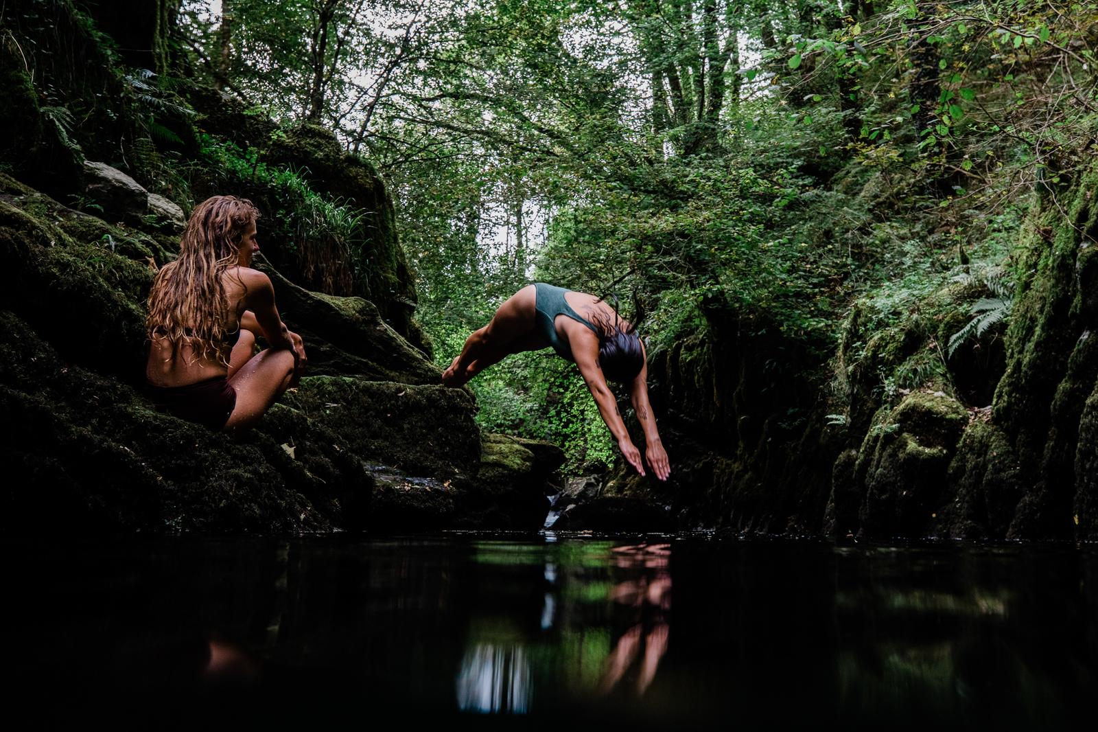 Two women in a forested area, one in a green swimsuit and one in a black swimsuit, both in a yoga pose near a body of water.