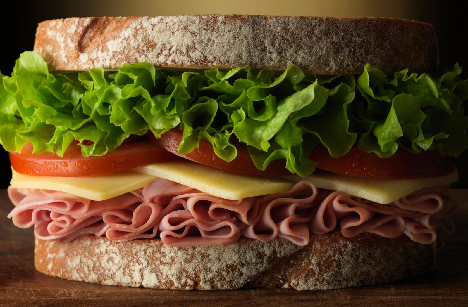 A close-up image of a large, layered sandwich with ham, cheese, tomato, and lettuce on a crusty bread.