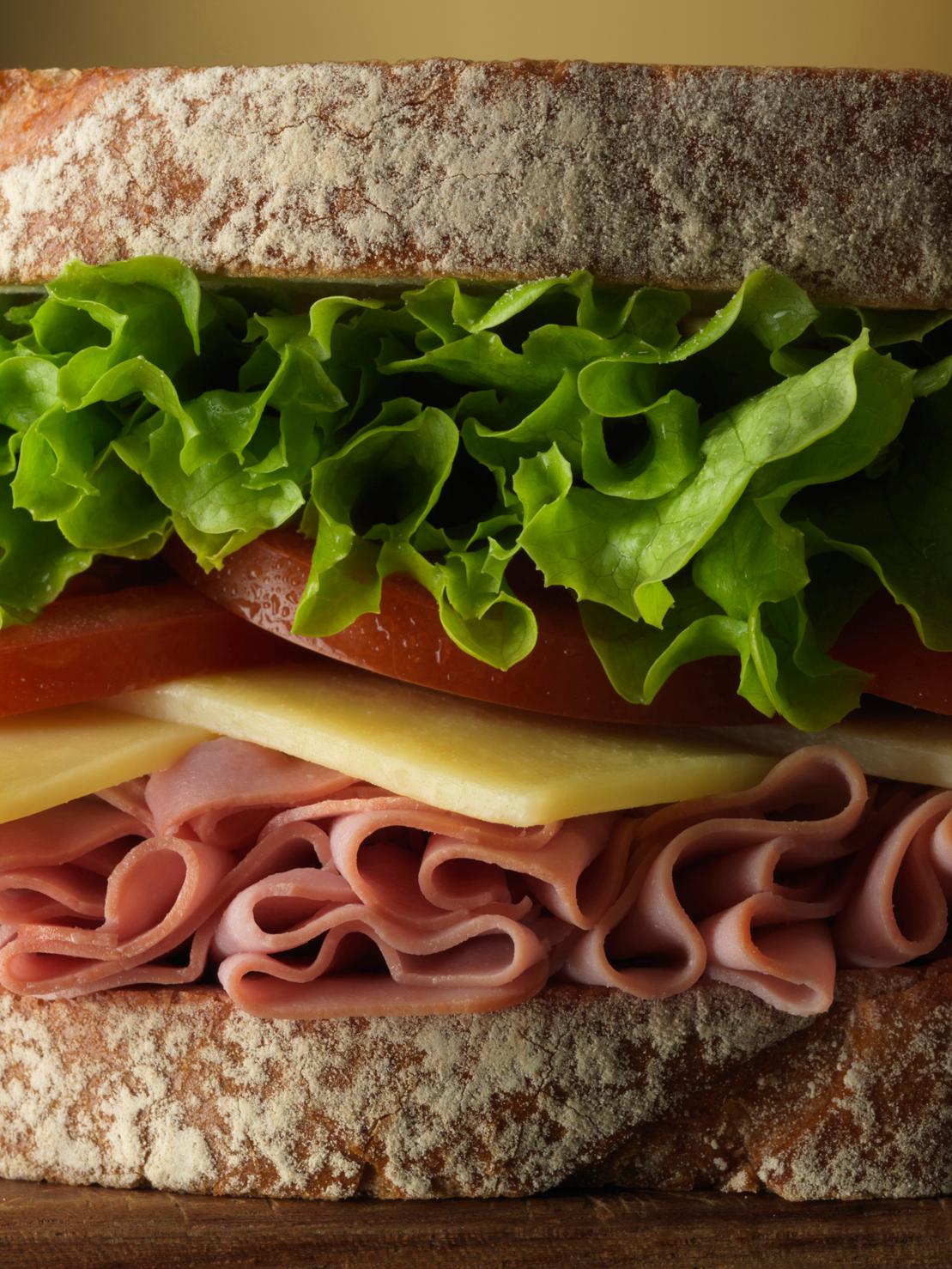 A close-up image of a large, layered sandwich with ham, cheese, tomato, and lettuce on a crusty bread.