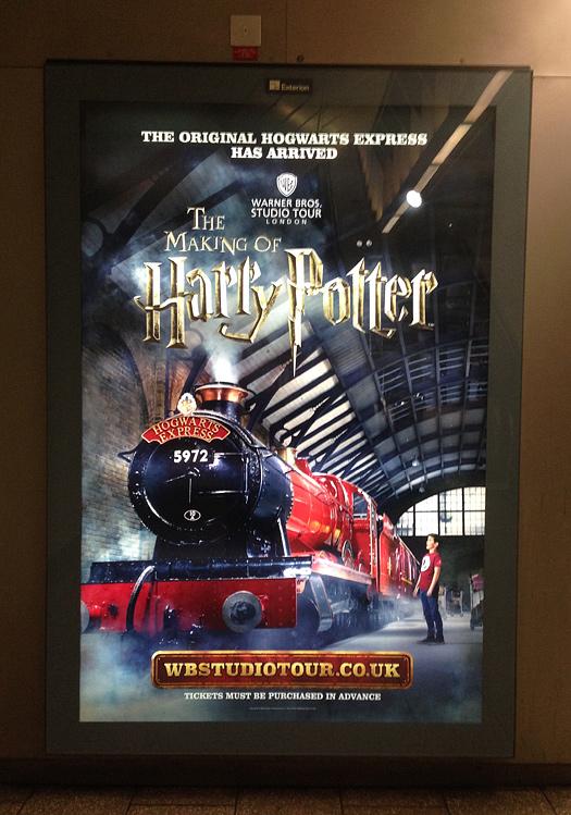 A red and black steam train, the Hogwarts Express, is seen in a station, with a large clock tower in the background.