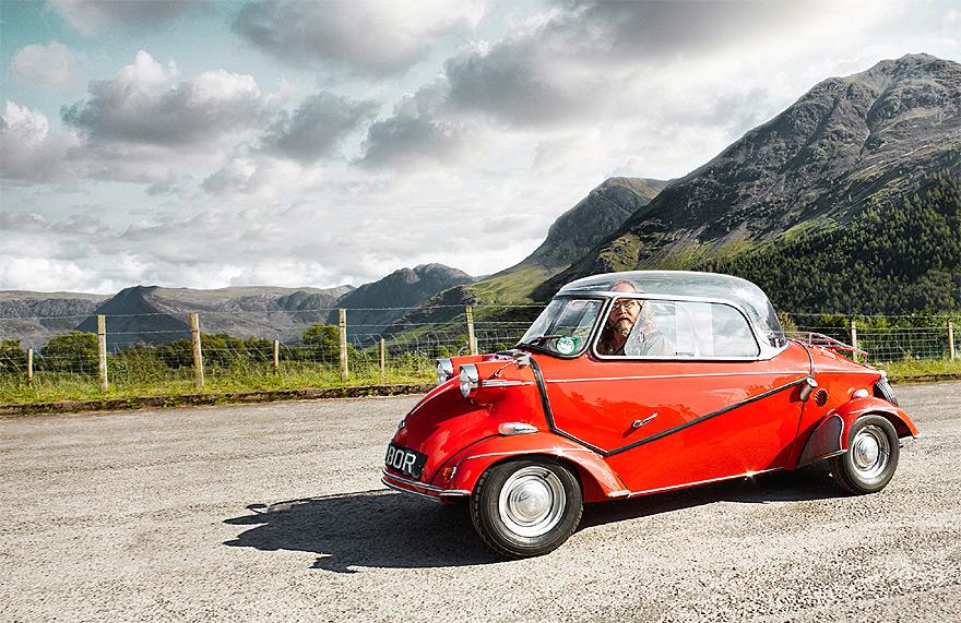 A red, vintage, two-seater car driving on a mountain road, with a mountain range in the background.