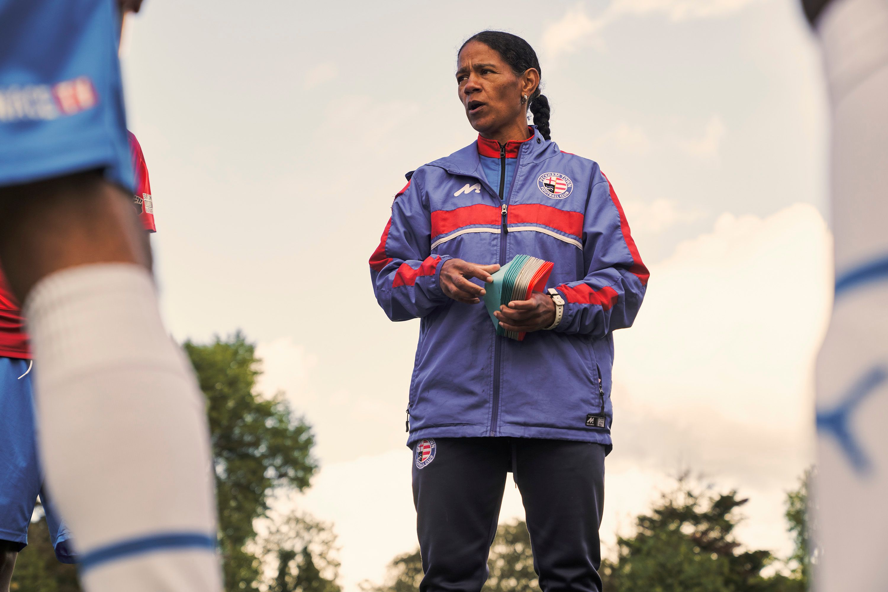 A woman wearing a blue and red Peckham Town Football Club jacket stands outdoors, holding a stack of colorful training markers while speaking to a group. Photograph by Nick Dolding.