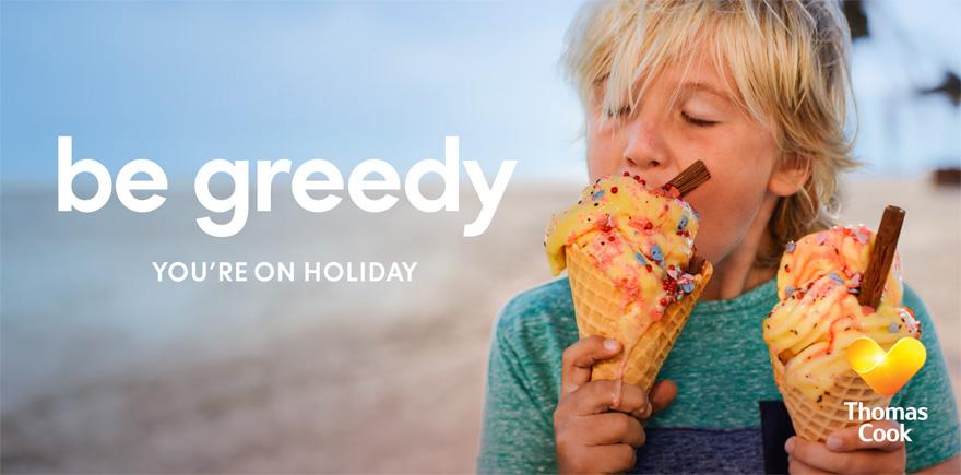 A young boy with blonde hair and a blue t-shirt is eating an ice cream cone on a beach.