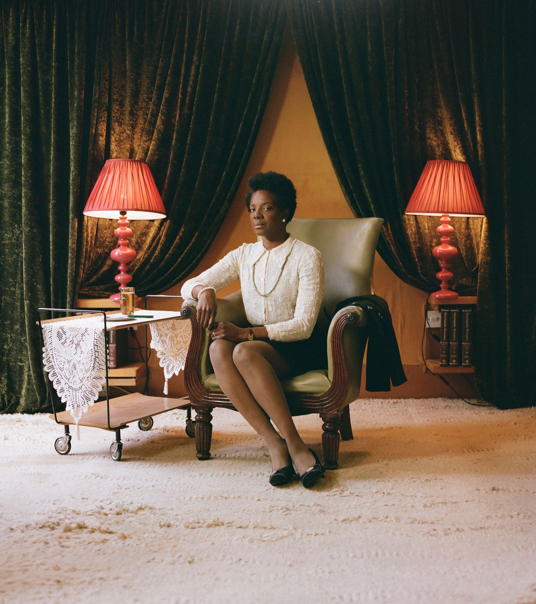 A woman with short, dark hair and a white, lace-trimmed blouse sits in a chair, with a small table to her left and a lamp behind her.