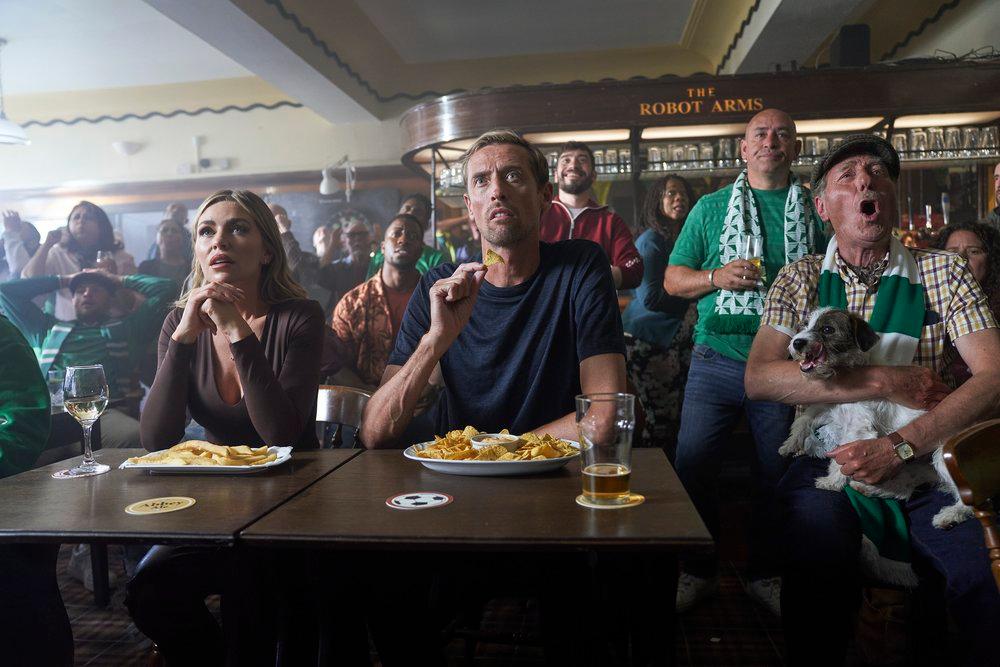 A crowded pub with people watching a football match, some wearing football jerseys, others holding dogs, and all enjoying snacks and drinks.