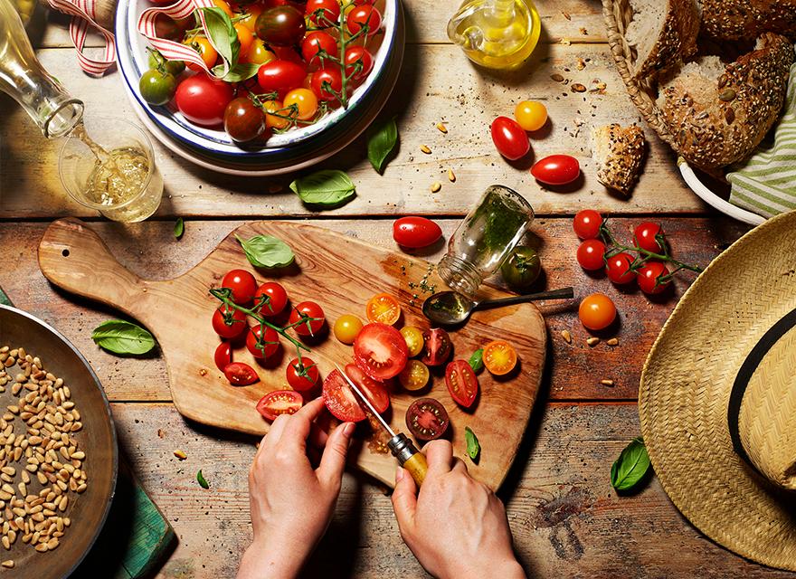 A person is chopping tomatoes on a wooden cutting board, surrounded by various food items and kitchen tools.
