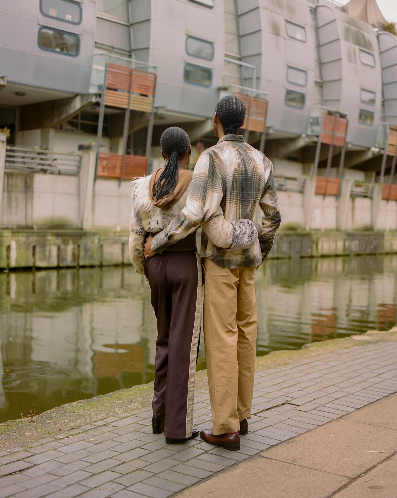 Two people standing back-to-back on a city street, arms around each other's waists, facing an industrial-style building.