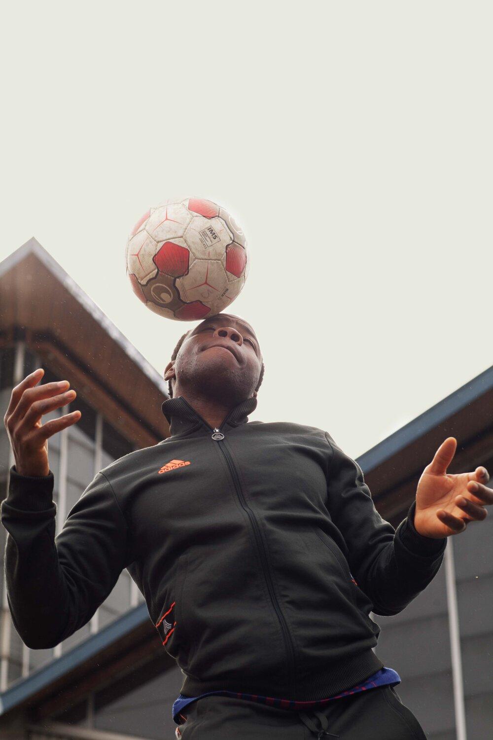 A person in a black Adidas jacket and black pants is balancing a soccer ball on their forehead, with their arms outstretched and their eyes closed.