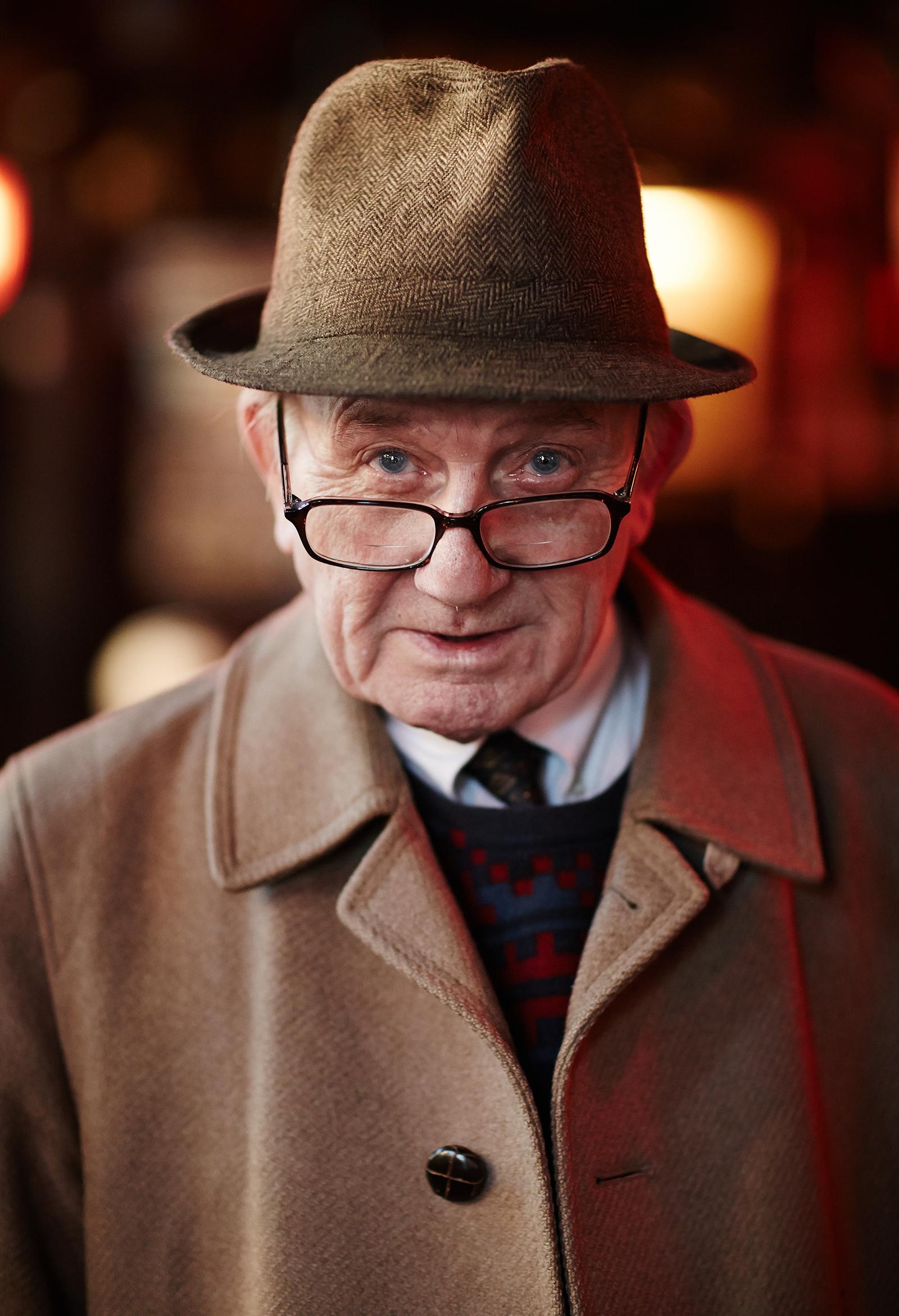 An elderly man wearing a brown fedora, a brown coat, a white shirt, a black tie, and a patterned sweater, with a blurred background.