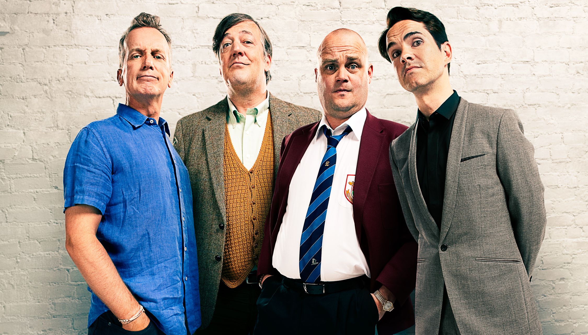 Three men in various outfits standing in front of a white brick wall, with one man in a blue shirt, one in a yellow sweater, and one in a red jacket.