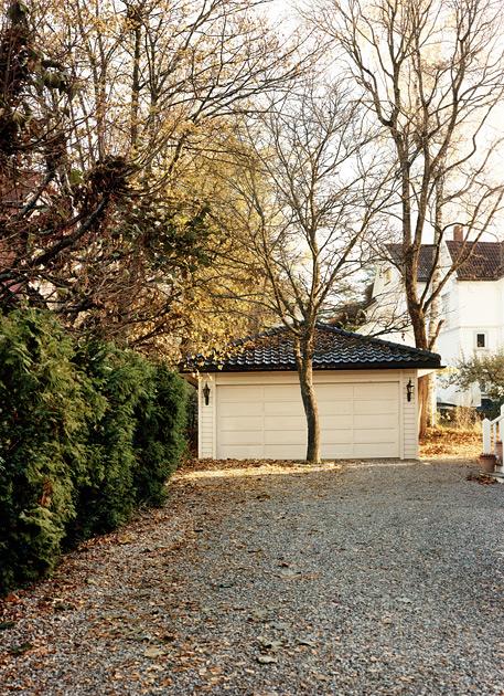 A white garage door with a black roof, surrounded by trees and a gravel driveway, with a house in the background.