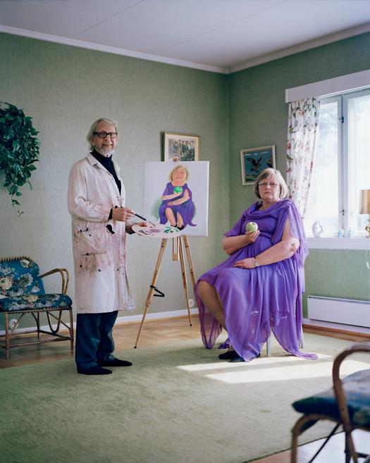 An artist in a studio, wearing a white painter's coat, standing next to an easel with a painting of a woman in a purple dress, holding a paintbrush and a palette.