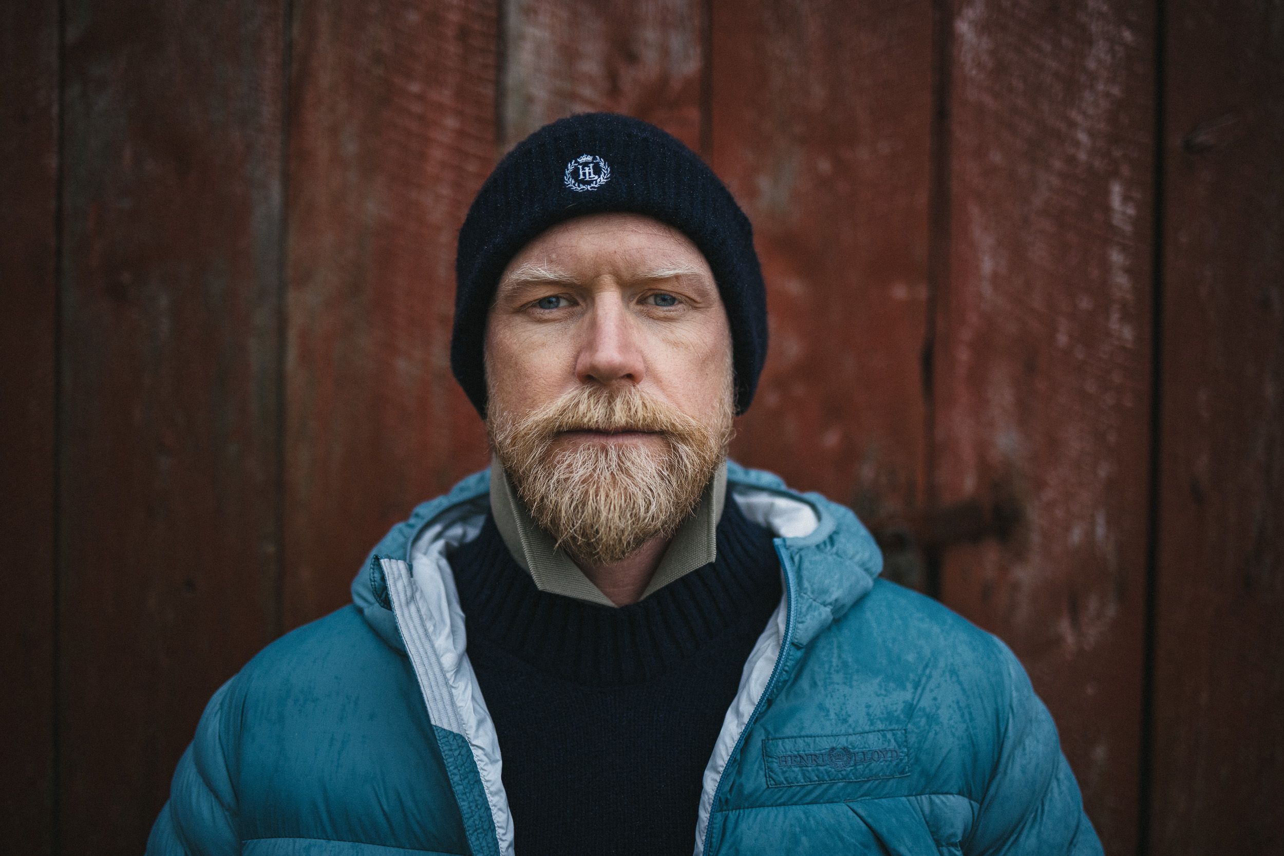 A man with a beard and a blue jacket standing in front of a wooden wall.