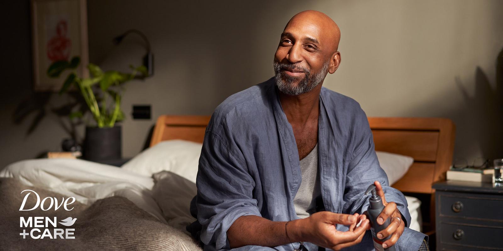 A man in a blue robe sits in a dimly lit room holding a bottle of fragrance.