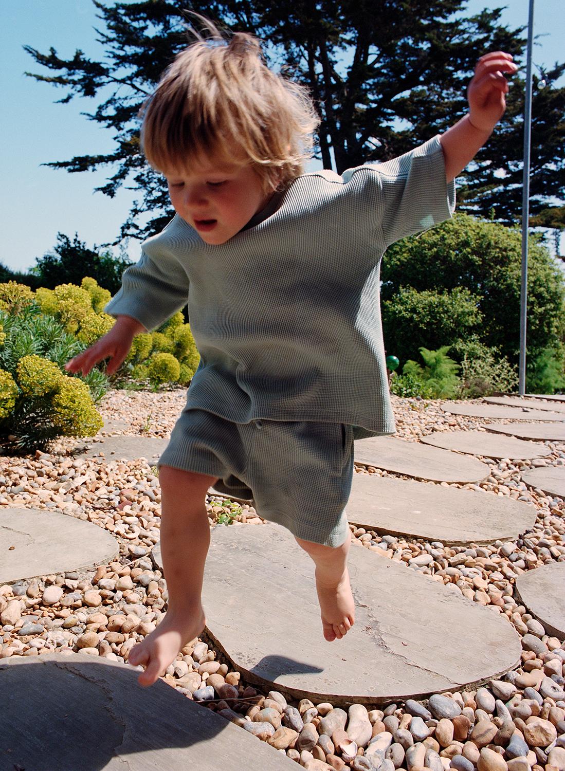 A young child in a green outfit is jumping on a large stone in a garden.