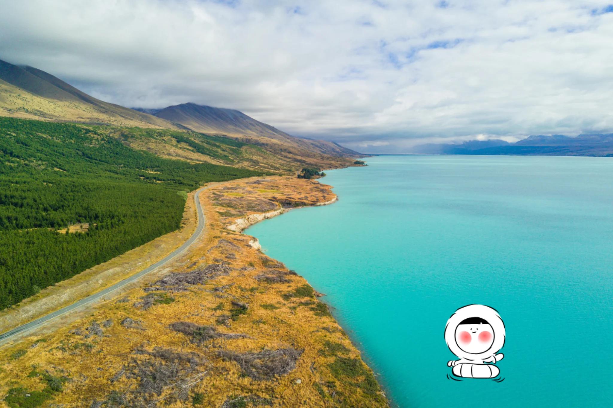 Long turquoise lake bordered by rolling hills and mountains, with a winding road along the shoreline.