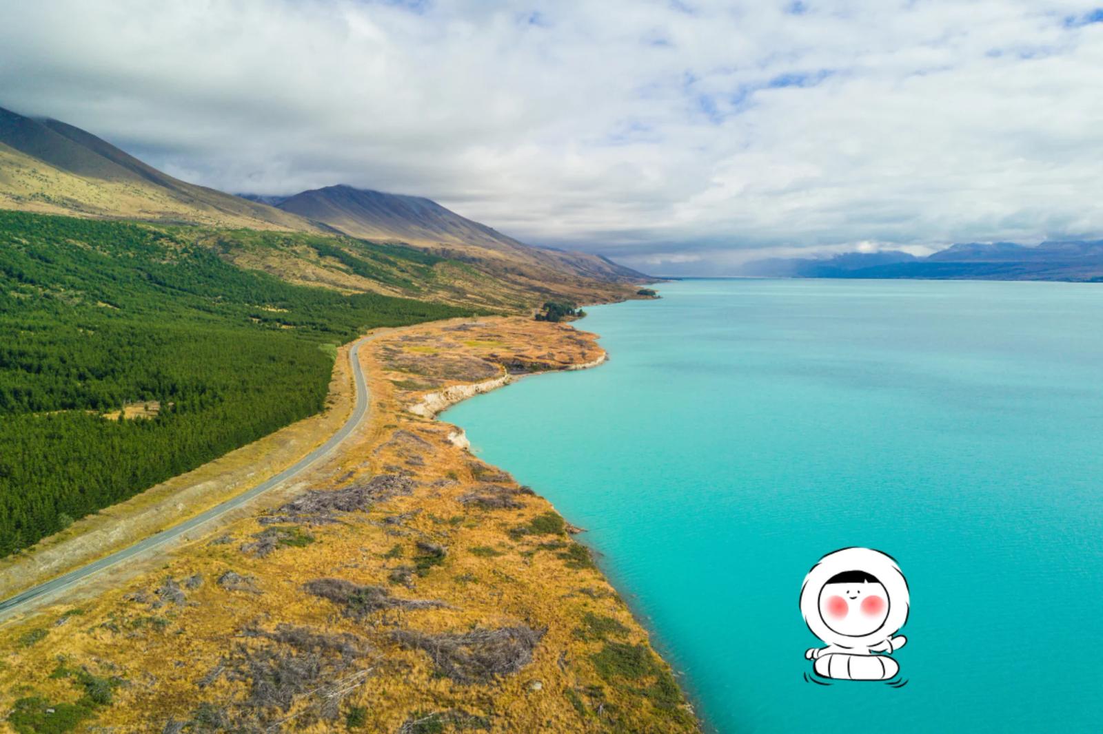 Long turquoise lake bordered by rolling hills and mountains, with a winding road along the shoreline.