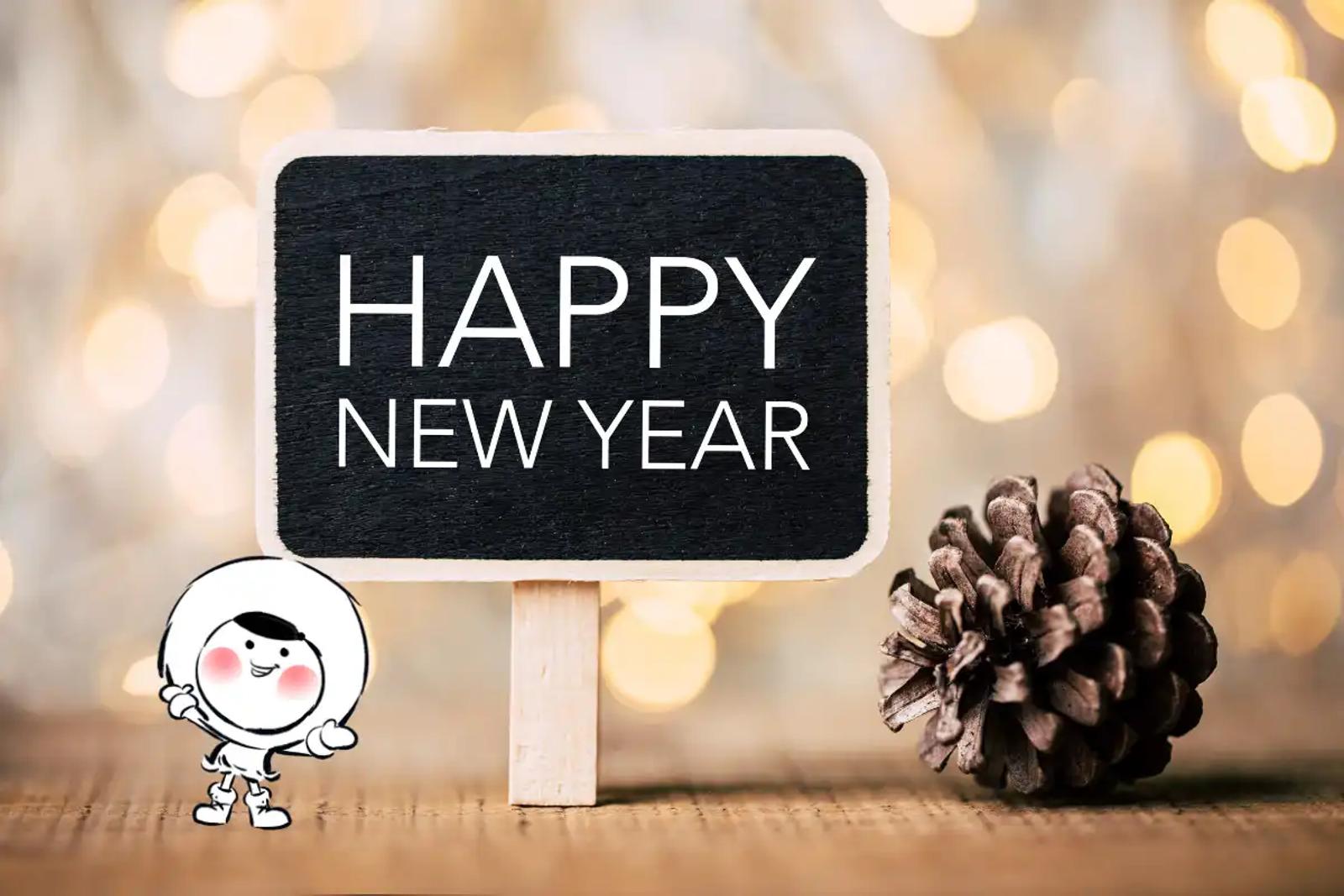 A small chalkboard sign with the words “HAPPY NEW YEAR” stands on a wooden surface next to a pinecone, with warm golden bokeh lights glowing softly in the background.
