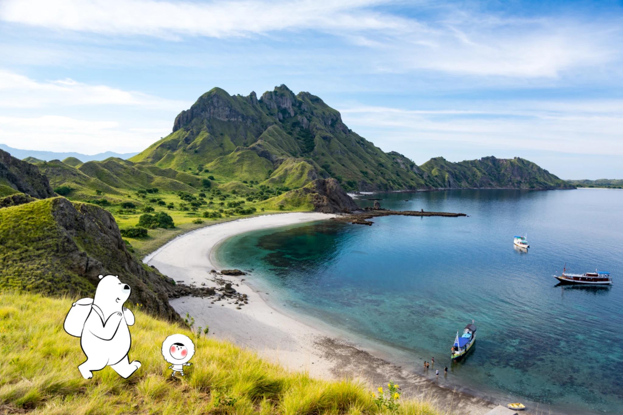 A sweeping coastal view of Komodo National Park featuring green hills, a crescent-shaped white sand beach, clear turquoise water, and several small boats anchored near the shore under a bright blue sky.