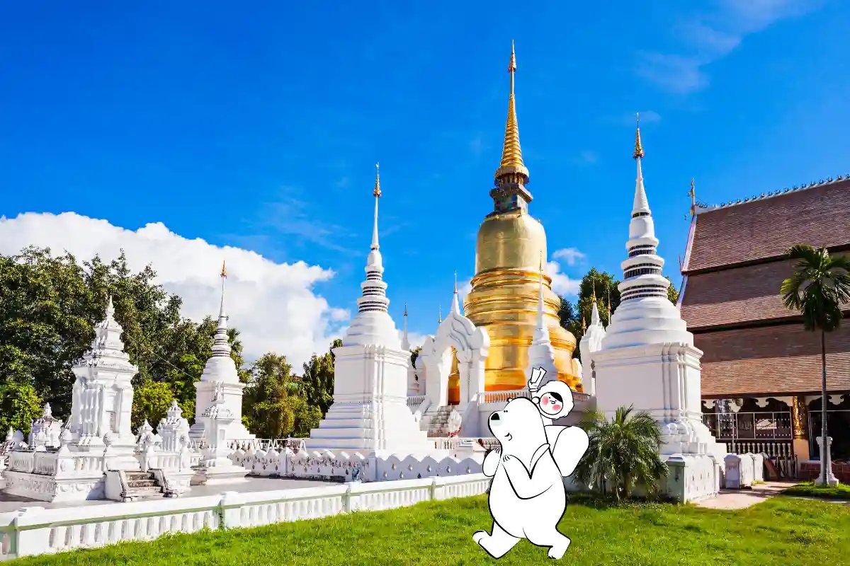 Golden stupa and white pagodas at a Chiang Mai temple under a bright blue sky, with Eskimo’s travel mascot walking peacefully on the grass.
