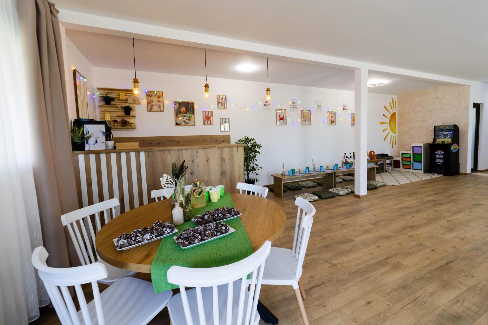 Masă rotundă cu prăjituri și limonadă, bar din lemn, ghirlande de lumini și colțul de joacă în fundal