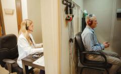 An audiologist at a laptop running a hearing test for a man wearing headphones in a sound booth