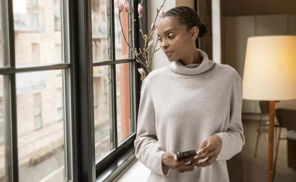 A woman in a gray sweater standing by a window, holding her phone