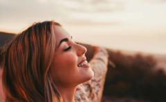 A smiling woman with her eyes closed and her arms open, facing a sunset sky