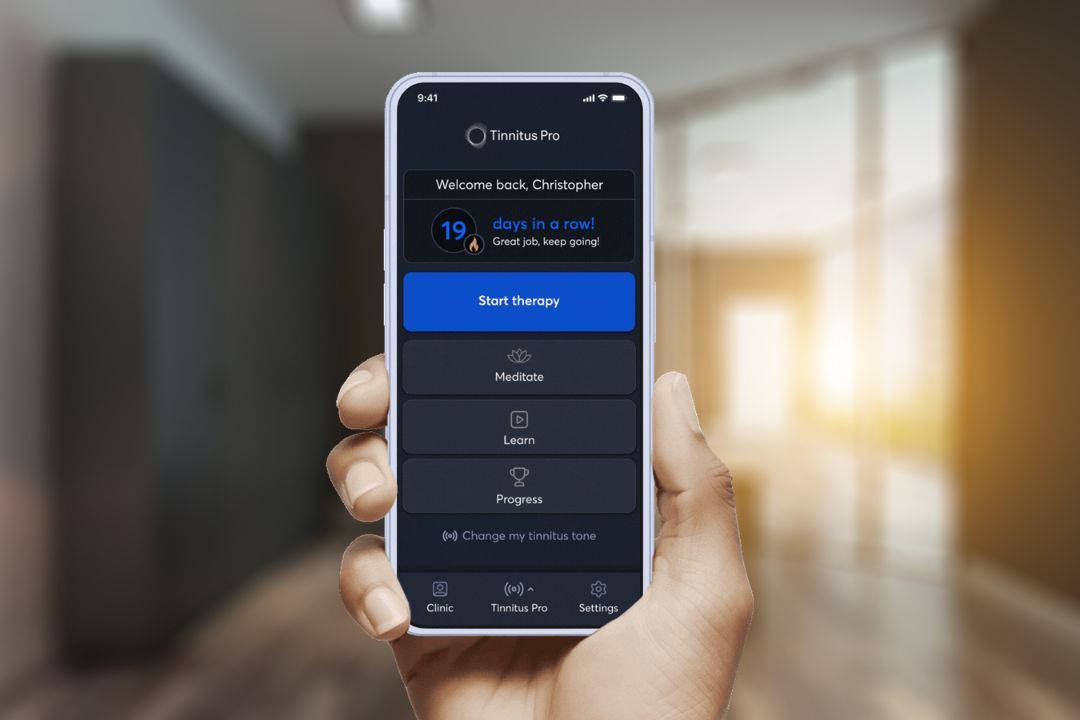 A hand holding a phone showing the Tinnitus Pro app, with options to start therapy, meditate, learn, and see progress