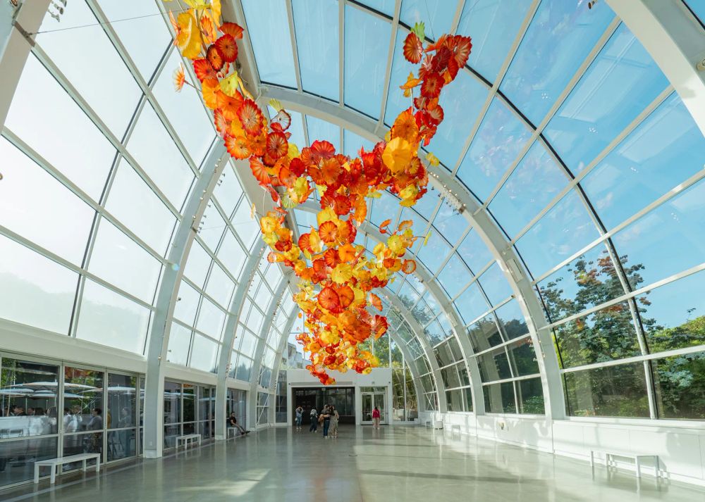 Orange suspended glass sculpture beneath the arched glazing of a bright glasshouse