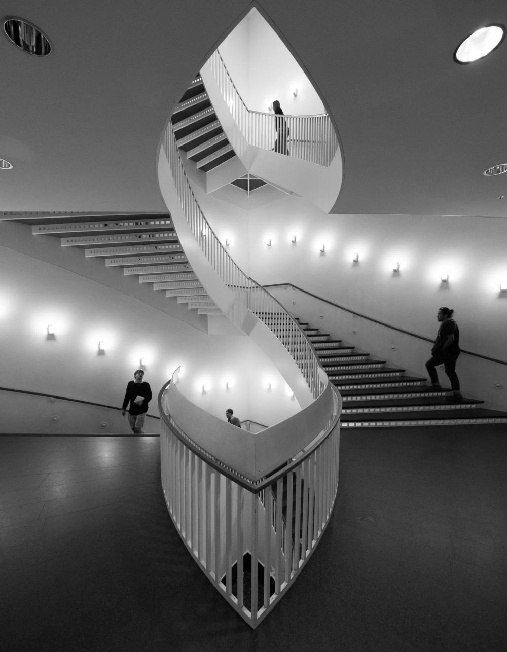 A twisting white stair with figures in a monochrome interior