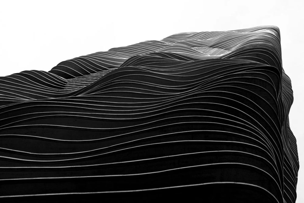 An undulating tower facade against a pale sky in black and white
