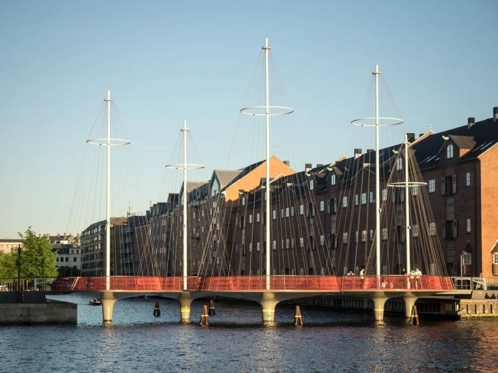 A slender red pedestrian bridge and white masts before waterfront buildings