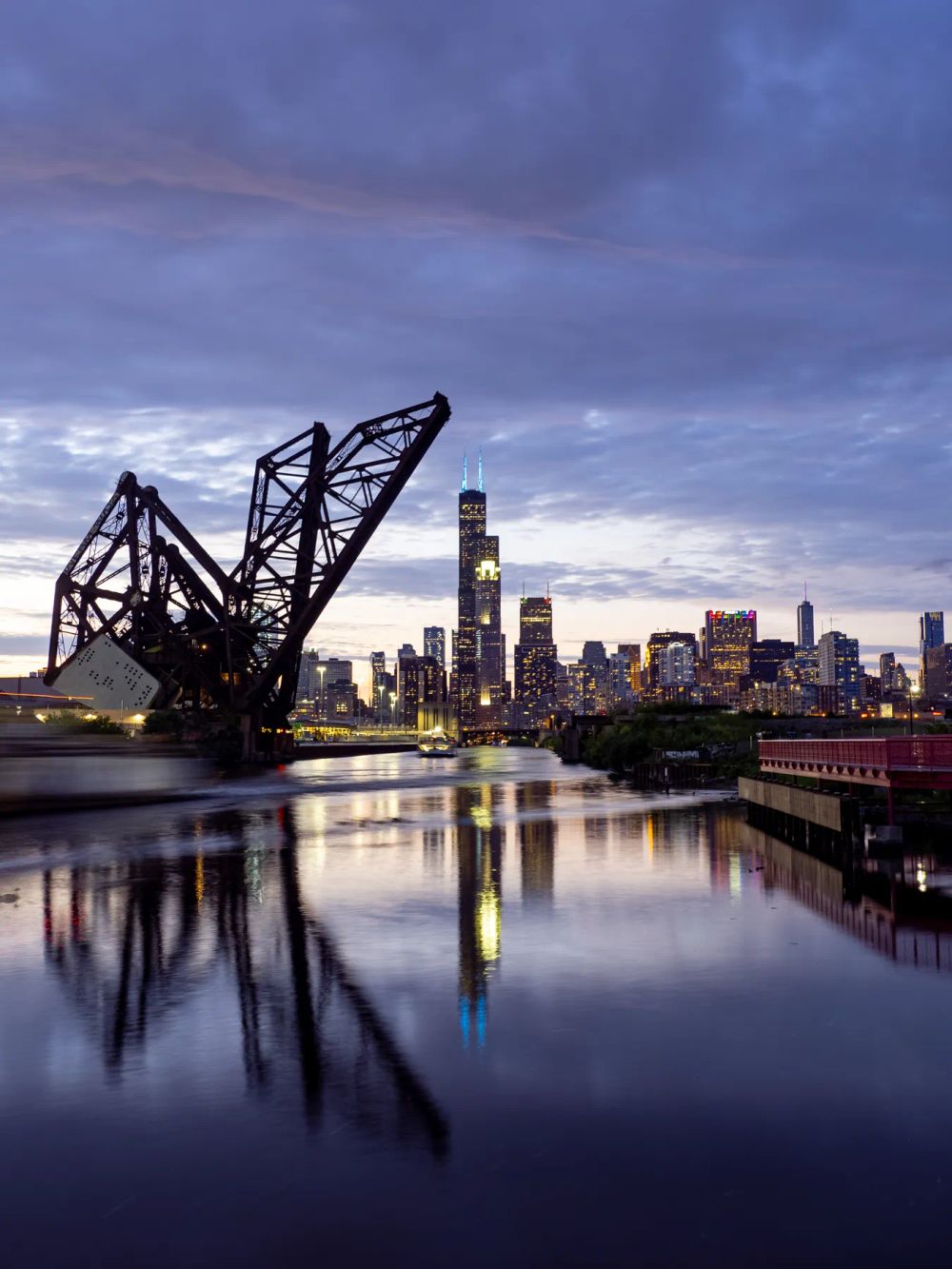 A raised steel bridge reflected in a river before a city skyline at dusk