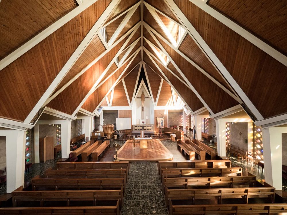 A timber church interior with triangular white roof trusses above wooden pews