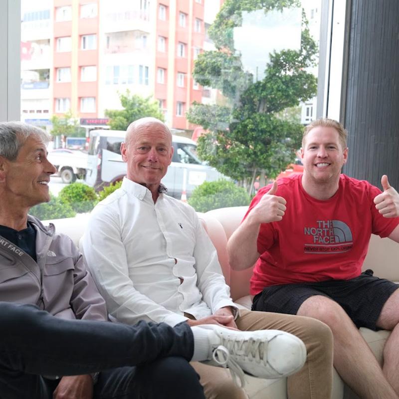 Three smiling men sitting on a sofa, two giving thumbs up