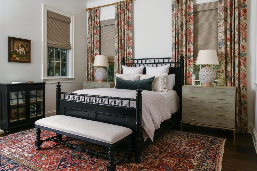Traditional guest bedroom with a black spindle bed frame, sage green dressers, floral curtains, and a red and blue Oriental rug