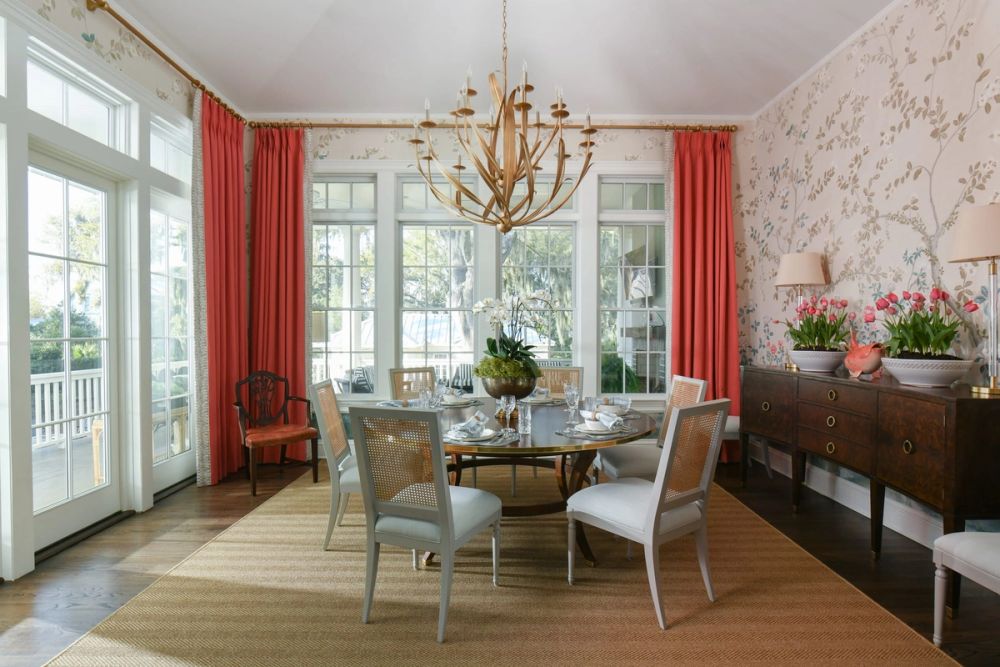 Elegant dining room with a round wood table, cane-backed chairs, gold chandelier, coral-pink curtains, and delicate floral wallpaper