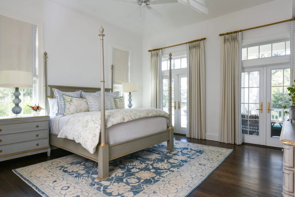 Primary bedroom featuring a grey four-poster bed with gold accents, blue and cream floral area rug, and cream curtains with blue geometric trim