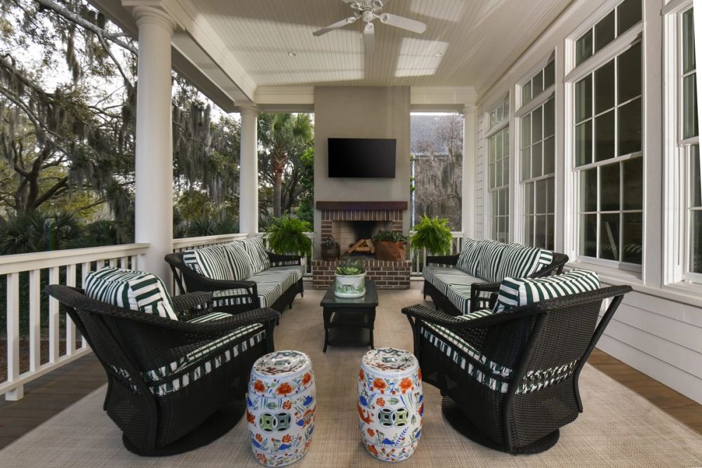 Covered outdoor porch with dark wicker seating, green and white striped cushions, brick fireplace, and views of moss-draped trees